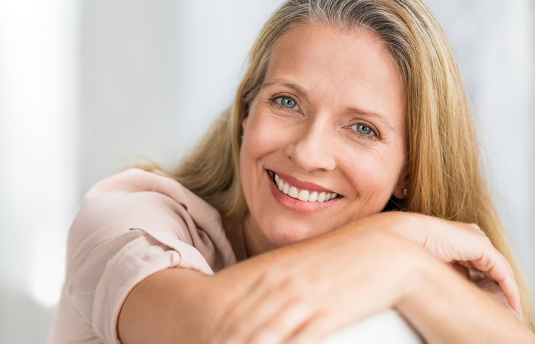 A smiling woman with long blonde hair rests her arms on a surface, looking at the camera. Martinez, GA