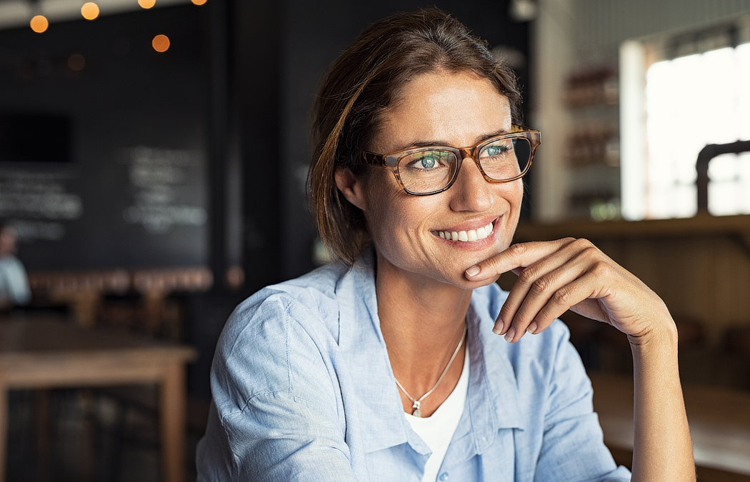 A woman wearing glasses and a light blue shirt smiles while resting her chin on her hand in a cafe setting. Martinez, GA