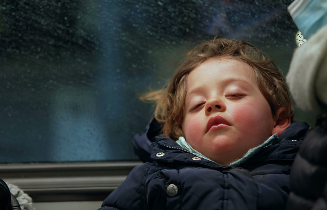 A child with closed eyes rests against a window on a rainy day. The child is wearing a dark jacket and a light scarf. Martinez, GA